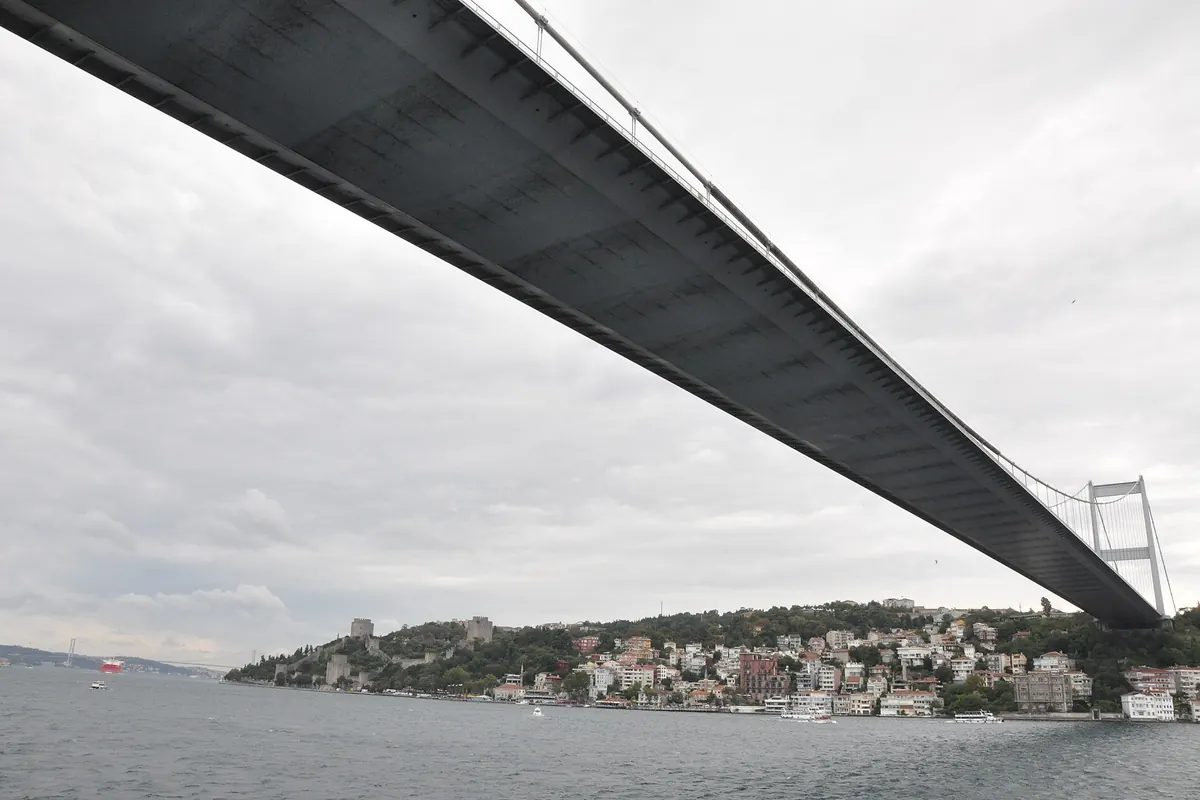 The underside of the Fatih Sultan Mehmet Bridge stretching across the strait, with the Rumeli Fortress on the wooded shore below.