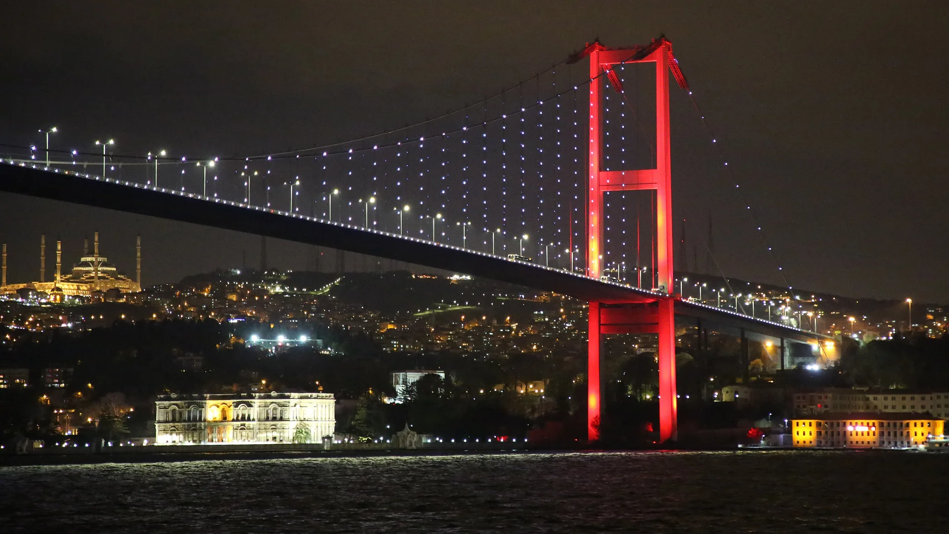 The Bosphorus Bridge at night with its tower lit red and its cables picked out in white lights, and a lit palace on the shore below it.